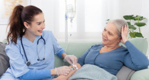 A young smiling nurse helps a sick elderly woman receive IV drip infusion and vitamin injection therapy while sitting on a sofa in a medical clinic or at home.
