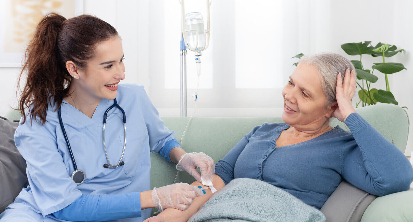 A young smiling nurse helps a sick elderly woman receive IV drip infusion and vitamin injection therapy while sitting on a sofa in a medical clinic or at home.